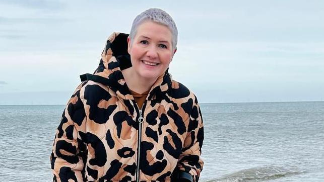 Angharad stood with the sea behind her. She has cropped white hair and is wearing a padded leopard print coat, and smiling at the camera.