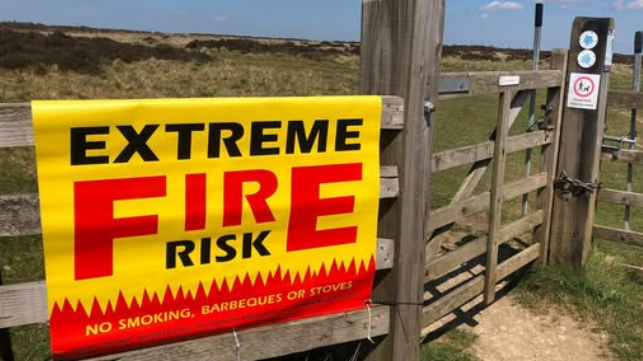 A sign - in vivid yellow and red - warning of the extreme risk of moorland fires. The sign is attached to a five-bar gate. Parched-looking moorland stretches away behind it.