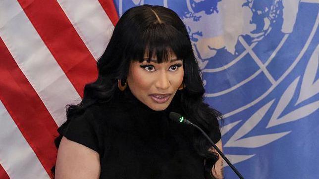 Nicki Minaj with long black hair and bangs, standing at a podium. She is dressed in a fitted black outfit. Behind her, two large flags hang side by side, one is the US  flag and the other has the UN emblem on a blue background