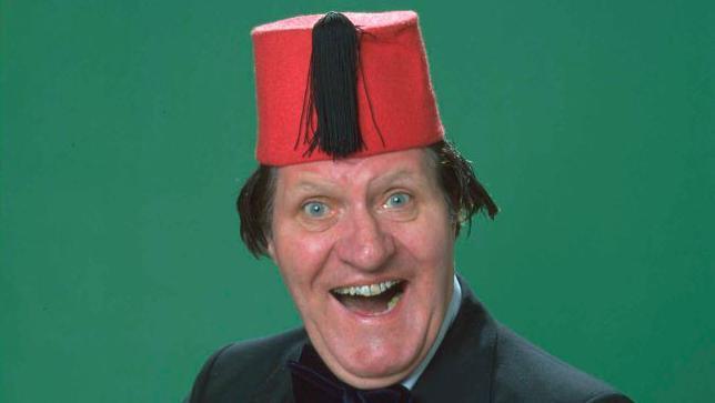 Tommy Cooper grins at the camera wearing a fez in front of a plain green background 