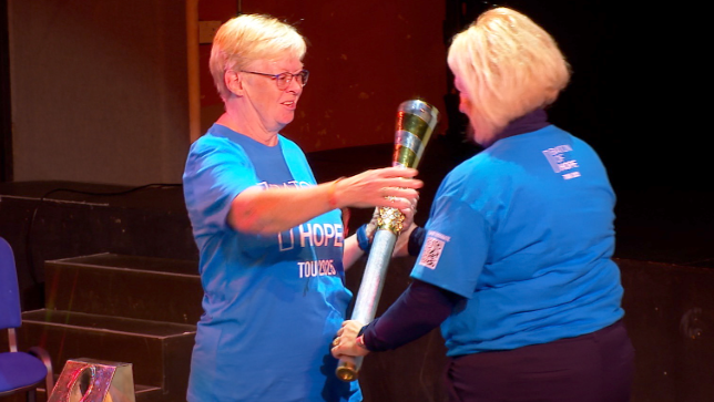 The image shows a ceremonial moment where two individuals in blue shirts are participating in a symbolic torch handover. One person is passing the torch with both hands, while the other is receiving it with one hand. The setting appears to be indoors, possibly on a stage, with chairs and equipment in the background.