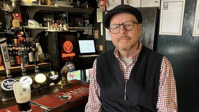 A man in a black cap, black gilet and red checked shirt is looking solemn while sitting at the bar of a traditional pub. 