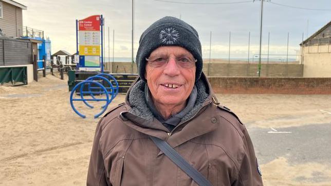 Peter Crome wears a brown jacket and stands on a beach 