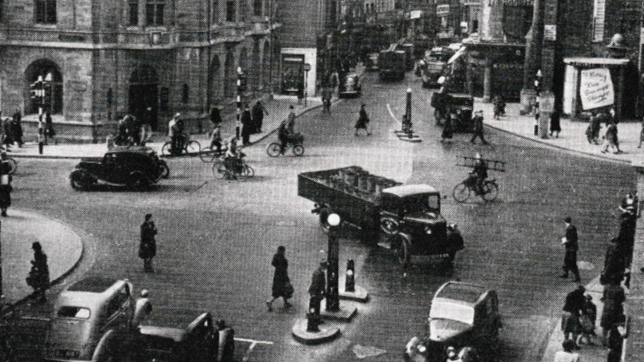 Carfax in Oxford city centre with vehicles and pedestrians. A cyclist is carrying a ladder as he rides from Cornmarket, across Carfax and to St Aldate's 