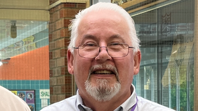 A close up of Andy Osborn. He wears rimmed glasses, has white hair and a short white beard. Behind him is a glass window