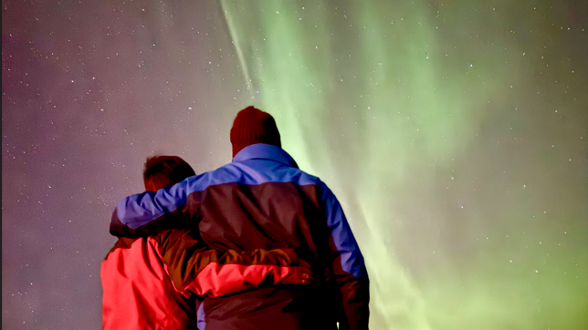 Two figures with backs to camera and arms around each other, one wearing a red waterproof jacket and other a black and blue waterproof jacket and black woolly hat, with the aurora of green lights in the sky in the background