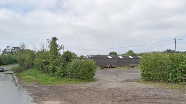 A country road runs alongside a farm gate entrance, between two hedgerows. The entrance is muddy, and beyond the gate there is an open gravel area with what looks old machinery and a barn. 