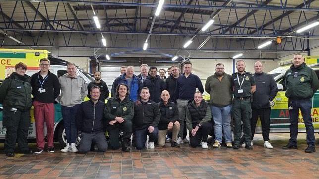 Newly-recruited volunteer responders attended an induction evening at Welwyn Garden City ambulance station in December, there are two rows of people, some crouching down, some standing, in a room with a tile floor, in a large room with ambulances in it. 