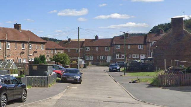 Brown brick houses in an Esh Winning street. There is a bike lying on its side and a number of parked cars. 