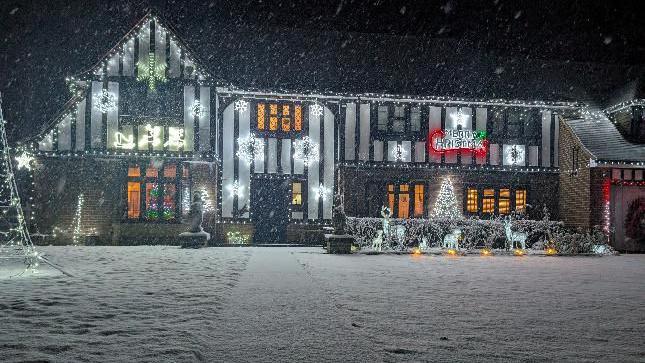 Snow falls outside a big white house, still lit up by Christmas lights