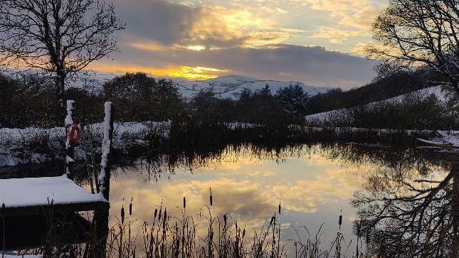 A sunset shines onto a pond, reflecting an orange sky and clouds. there is a dock covered in snow and snow around the banks of the pond on mountains in the distance