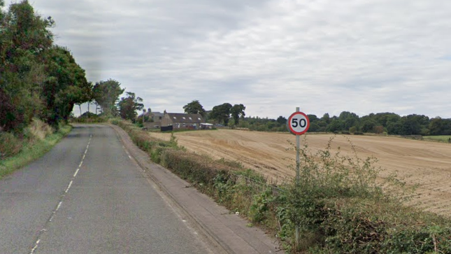 A road bordered by hedges and fields with a 50mph sign at the edge. A house can be seen in the distance.