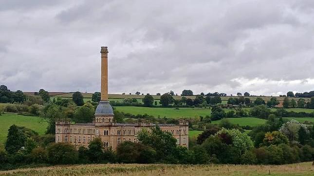 Chipping Norton's Bliss Tweed Mill - a pale stone building with a tall chimney - in the foreground and to the left of the frame. The mill's chimney reaches high into the sky of grey clouds. There are rolling green fields, hedges and trees in the background