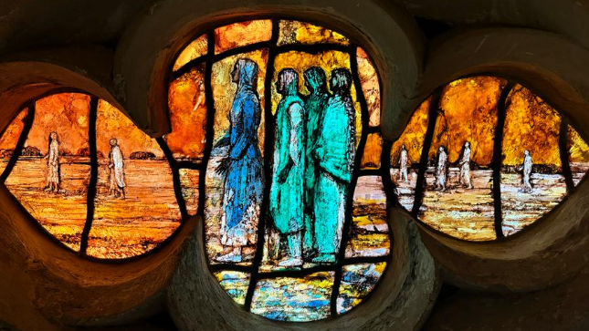 A close-up of a pane of the glass in a flower shape with several biblical figures depicted in hues of orange, turquoise and blue