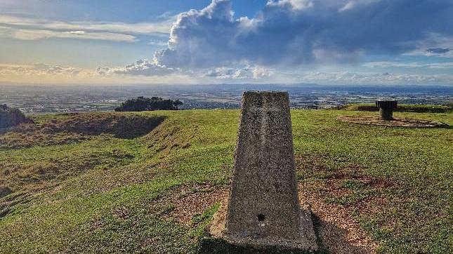 A landmark stone, almost like a grave with a cross lightly etched into it, stands on top of a very large hill overlooking a valley. There is a large cloud rolling across the sky and the sunlight is golden, at what appears to be just before sunset.