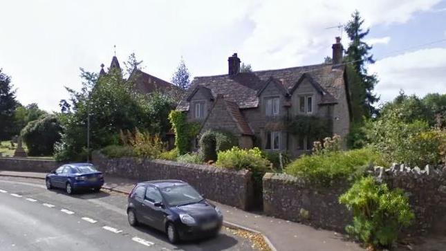 Church Cottage - a brick building surrounded with trees and bushes, with a stone wall at the front.