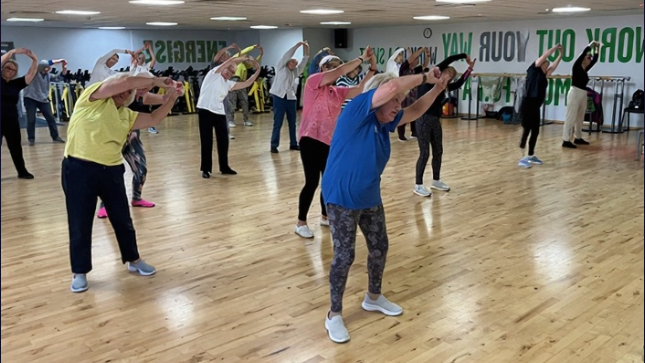 Dance and fitness instructor Maureen Conroy leads a class in a large gym. She holds her hands above her head as her students - behind her - follow her movements. The gym has a polished wooden floor.