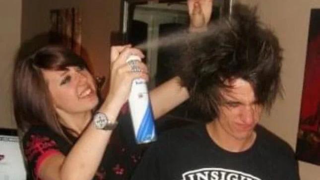 A girl with brown hair and a fringe using hair spray on a boy with long brown hair. She is styling his hair to stay up. 