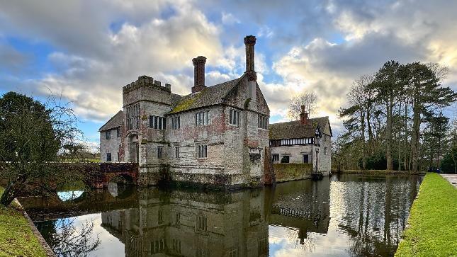 An old stone-walled building with tall chimneys, a gatehouse and a bridge sitting in the middle of a small lake with grass surrounding it