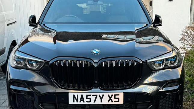 A black BMW X5 on a drive. It is in clean condition and has a customise number plate which reads NA57Y X5. There is a van parked next to it. 