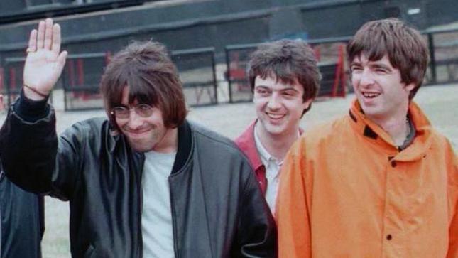 Oasis offstage at their Knebworth shows in 1996 - Liam in tinted glasses raising a palm, with Noel Gallagher next to him and Paul McGuigan behind them