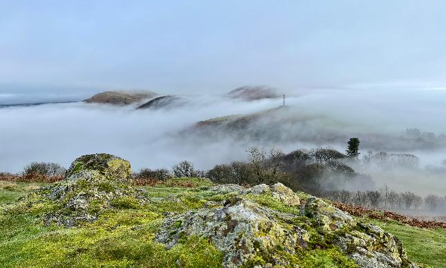 Cloud and mist can be seen blanketing hills in Church Stretton. Several hills are seen emerging from mist and fogs with some trees.