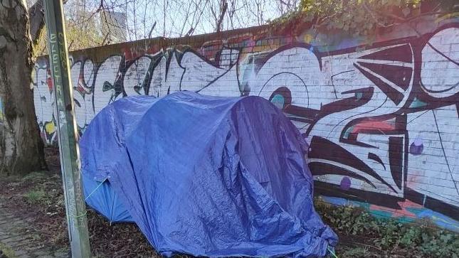 Blue tent at pitched up by a lamppost with a wall full of graffiti in the background.