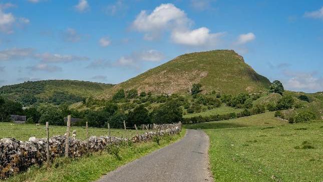 A country road in sunshine with a stone wall running along one side leading to a grassy hill in the distance with grass on either sides of the road and trees around the base of the hill