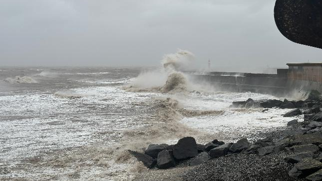 The seafront in Watchet, Somerset. Big waves can be seen crashing into the sea wall. The sky is very grey, cloudy and bleak. 