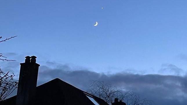 The crescent moon and Venus seen above a house roof against a light blue sky