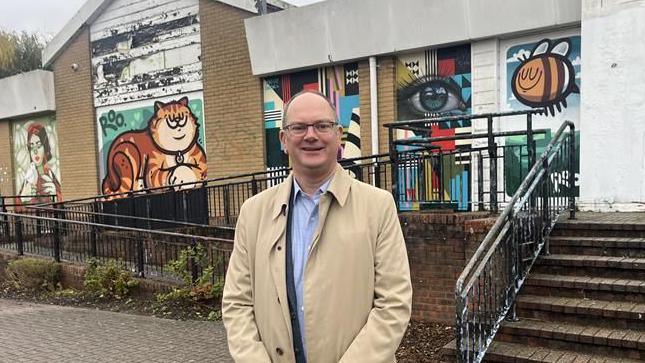 A bald man wearing a light blue shirt and beige jacket. He is stood outside a boarded-up, single storey building covered in various murals. He is looking into the camera and smiling
