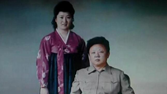Ko Yong Hui, in a purple and green Korean dress stands on the left. Kim Jong Il sits besides her