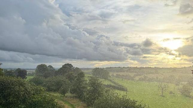 A Turneresque sky of hazy heather skies, filled with fluffy clouds and soft, golden sunlight to the far right. Below are rolling fields of various shades of green with trees and a pathway to the left of the foreground and more trees and hedgerows on the horizon