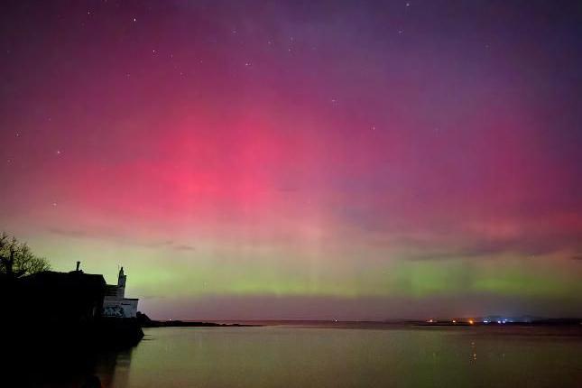 Pink and green lights in the sky with a house on the left and the lights from buildings just visible on the right.