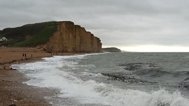 Waves rolling in at West Bay in the foreground, with the Jurassic Coast cliffs in the background and to the left of the image. There are a few people on the beach, close to the sea, just before the cliffs. The sea is dark with white foam and the sky above is grey. The cliff sides and beach are brown and there is grass on top of the cliffs.