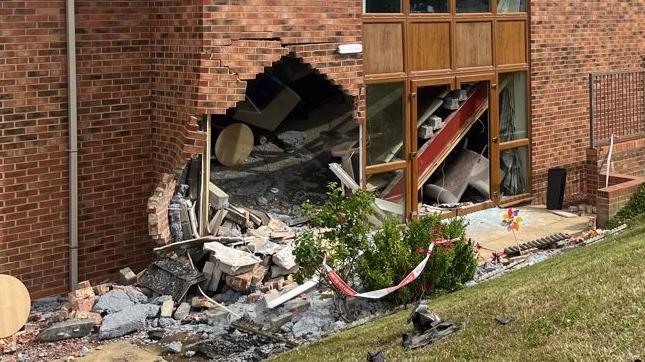 A large hole at the bottom left of a brick and white stone building next to a window. Bricks and debris from the wall are scattered all around and in the bushes and grass in front of the building. 