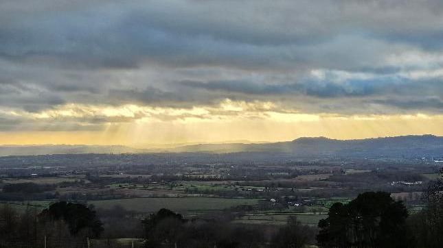 Yellow sun breaking through thick cloud over a dark landscape with fields and scattered houses.