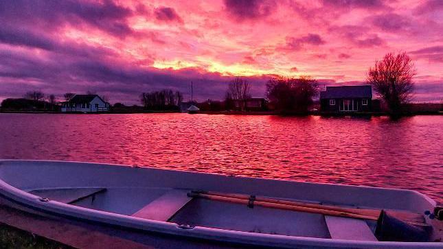 A rowing boat in the foreground on the edge of the River Thurne. Riverside homes on the other side of the river and the sky is a combination of purple, orange and red