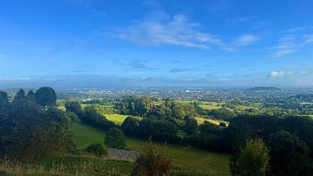 The view across a valley, the fields in the foreground are in the shade while the urban areas in the back are illuminated by sunshine. There is very little cloud in the bright blue sky.