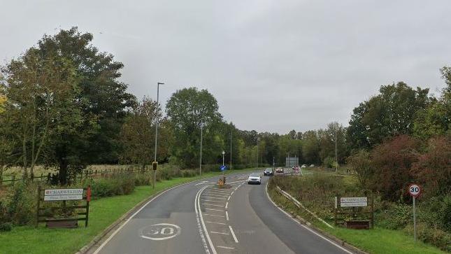 Road entrance to village, with Charwelton signs on either side of the carriageway. There is also a small 30 MPH sign to the right.  A right filter visible in the distance. There are hashes in the centre of the carriageway and a bollard. The road is lined with hedges and trees.