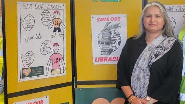 A woman pictured in front of a yellow display board with children's Save Our Library posters pinned on it. She has shoulder-length white grey hair and is wearing a grey floral scarf over a black top.