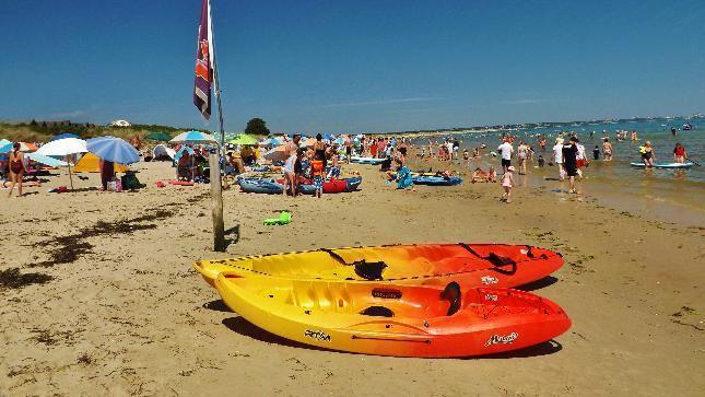 A busy sandy beach filled with people on the shore and in the sea. In the foreground are two orange and yellow kayaks