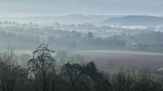 Mist covered fields with a ploughed field surrounded by trees, becoming more indistinct in the background.