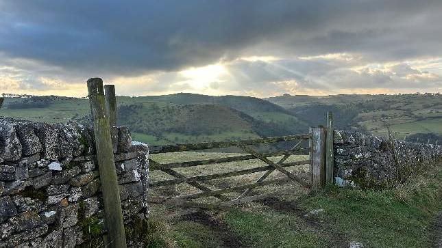 A wooden gate in a stone wall with a muddy track running through it and grassy hills in the distance and the sunlight breaking through the cloud overhead