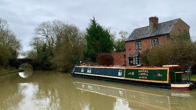 A dark green narrowboat on a green canal in front of a red brick house with a black roof and there is a small bridge in the distance.