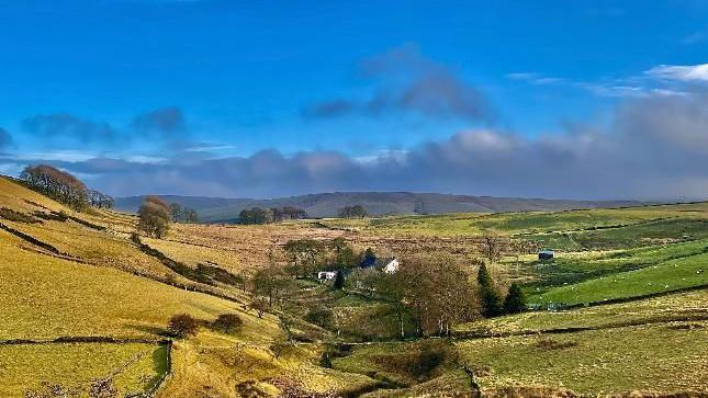 Rolling fields lead up from a shallow valley with blue sky overhead and some low-lying, wispy clouds.