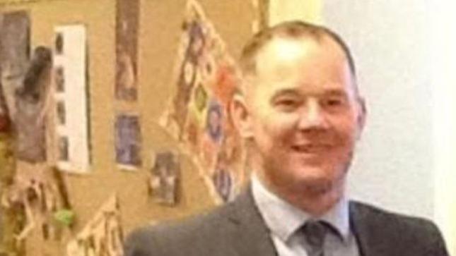 A picture of James 'Jamie' Richards in what appears to be a school environment, wearing a grey suit and tie. He is smiling and looks off from the camera. 