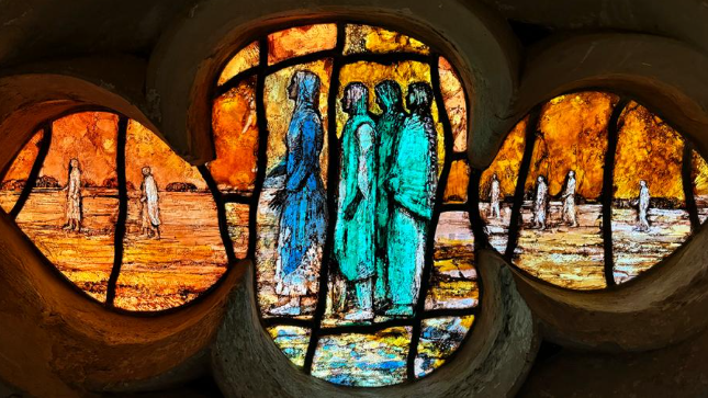 A close-up of a pane of the glass in a flower shape with several biblical figures depicted in hues of orange, turquoise and blue