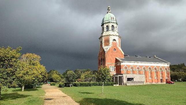 The Royal Victoria Hospital Chapel  - to the right of the shot - lit up by the sun but under dark, ominous clouds. There is a lawn in the foreground with a path leading to the chapel, with trees to the left of the frame and in front of the chapel, which is sideways on to the camera