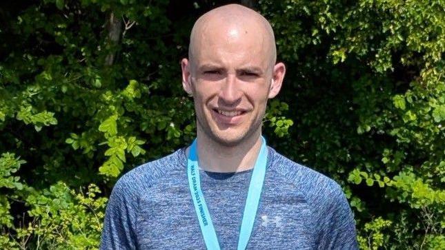 Thomas Mills wears a blue top with a blue ribbon of a medal around his neck. The ribbon says "HALF DRAM 2025 FINISHER". He smiles and stands in front of a tree with lots of green leaves.